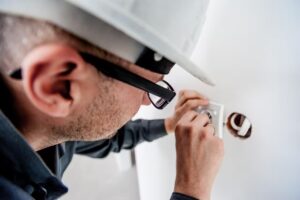 Électricien portant un casque de sécurité blanc et des lunettes, en train de réparer ou d’installer une prise électrique murale à l’aide d’un tournevis, avec des fils électriques apparents dans le mur.